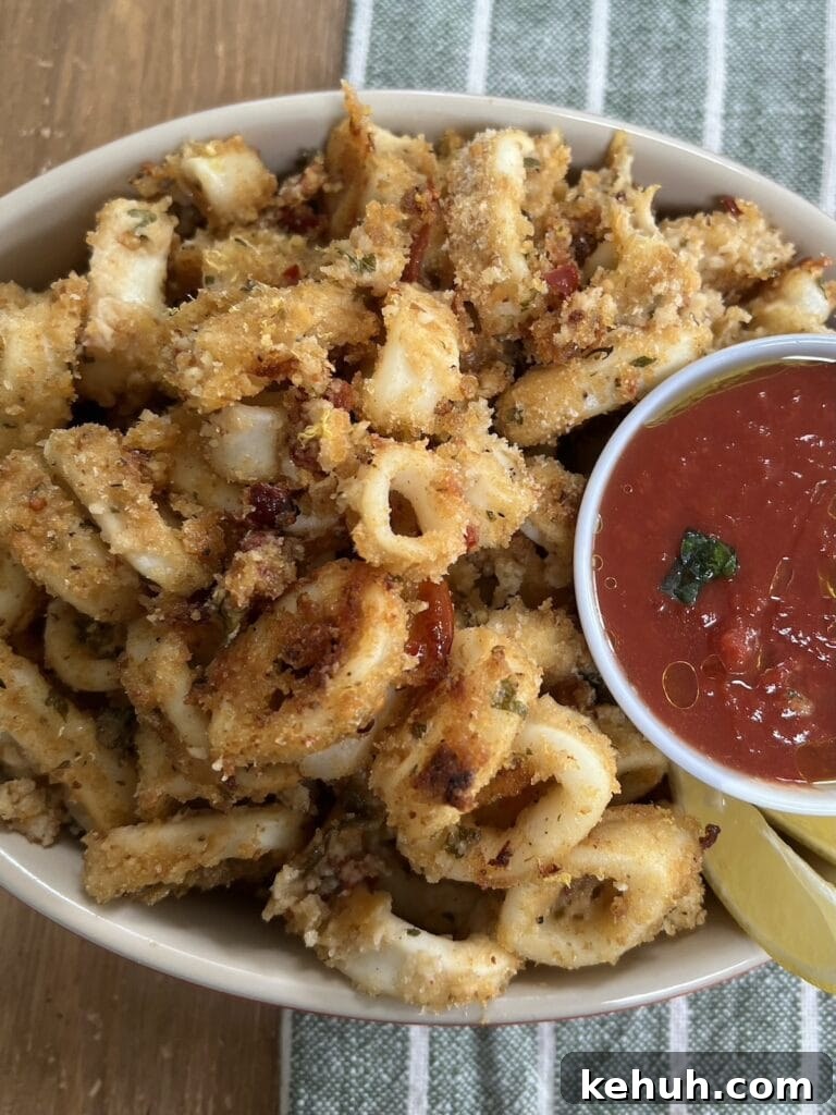 Crispy Baked Calamari: Oven-Fried Taste Without Deep Frying 9 Crispy Baked Calamari with small ramekin of marinara dipping sauce abd lemon wedges on white plate over green decorative towel.