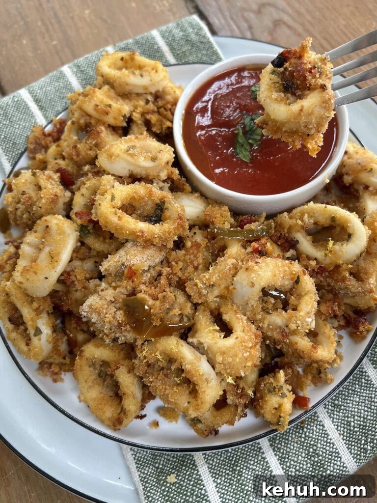 Crispy Baked Calamari: Oven-Fried Taste Without Deep Frying 10 Dipping a piece of oven fried and baked calamari in tomato sauce with fork next to whole plate of it.