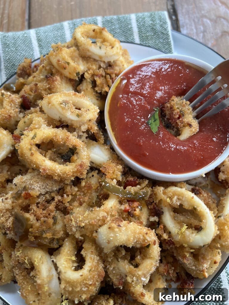 Crispy Baked Calamari: Oven-Fried Taste Without Deep Frying 3 Dipping a piece of oven fried and baked calamari in tomato sauce with fork next to whole plate of it.