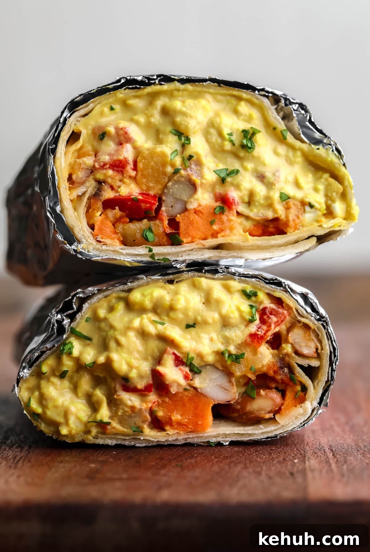 High-Protein Vegan Breakfast Burritos for Busy Mornings 2 burrito cut in half with foil around it on wood board with eggy mixture plus veggies