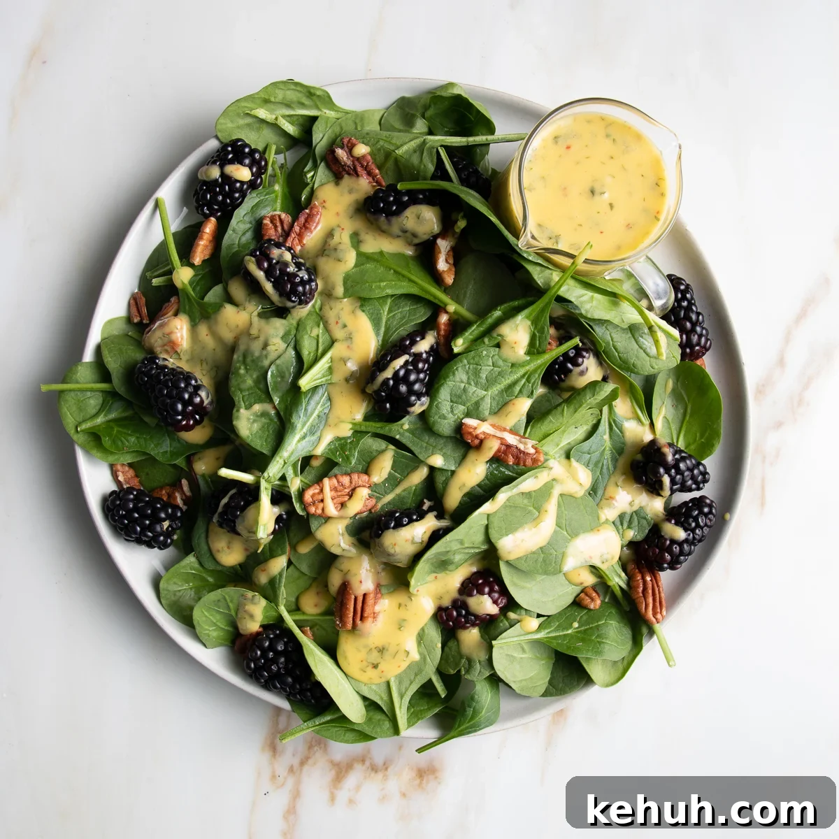 Vibrant Vegan Mango Salad Dressing Recipe for Fresh Salads 14 Spinach salad with blackberries, pecans and drizzled with mango dressing. There is a small glass pitcher with extra yellow, mango dressing on the side of the round white rimmed plate.