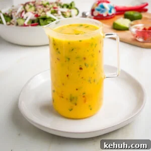 Vibrant Vegan Mango Salad Dressing Recipe for Fresh Salads 17 Small glass pitcher filled with bright yellow salad dressing with specks of red and green. In the background is a bowl of salad and some squeezed limes.