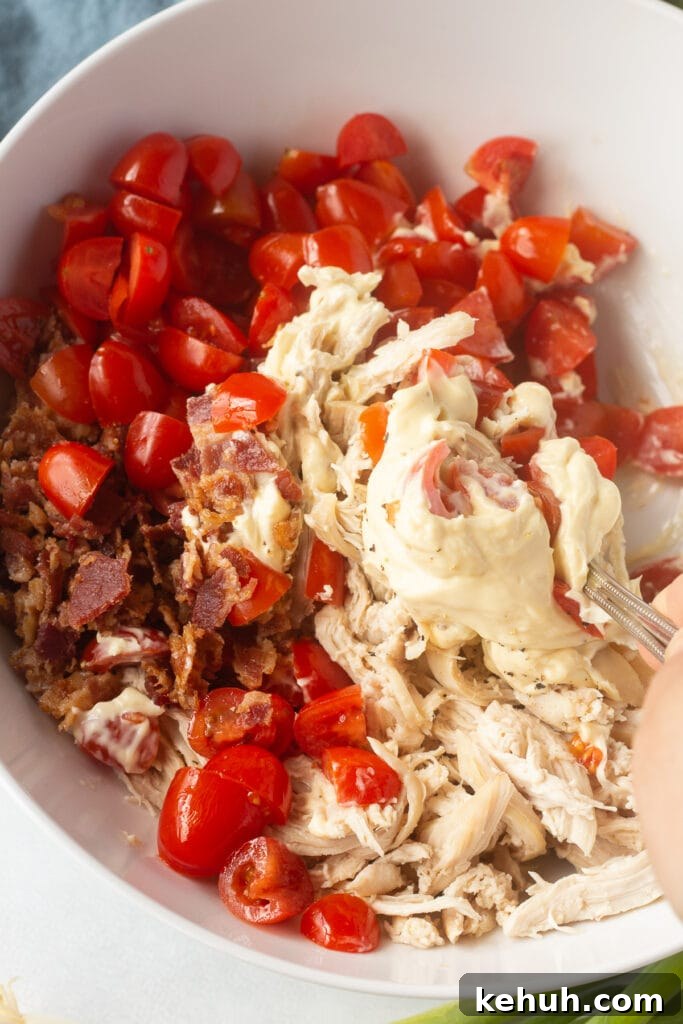 5-Ingredient BLT Chicken Salad Recipe for Quick Lunch 12 A spoon mixing mayo, bacon, chicken, pepper, and tomatoes together in a white bowl.