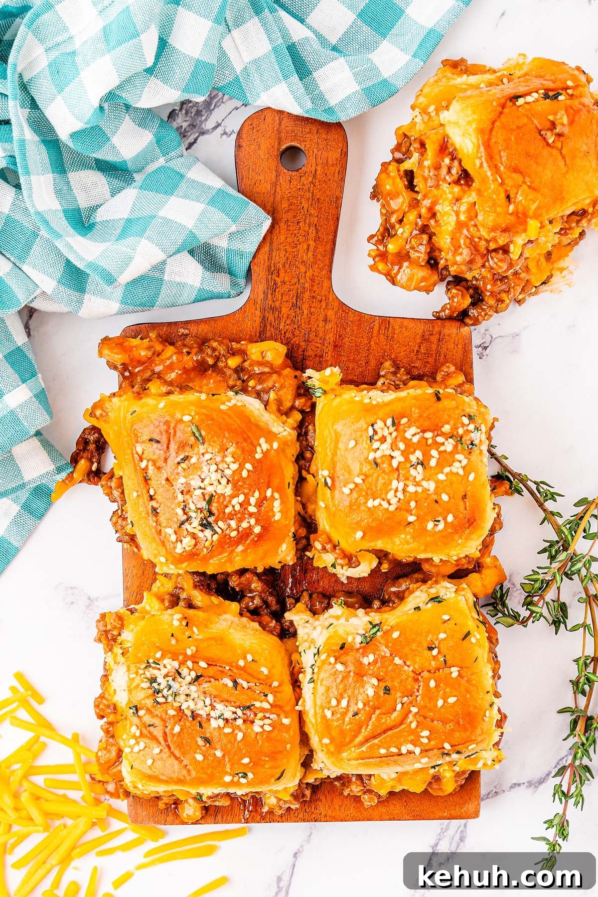 Savory Sloppy Joe Sliders Recipe for Game Day and Parties 11 Four baked sloppy joe sliders with beef and cheese on sesame seed buns, presented on a wooden board and garnished with herbs. Shredded cheese and a checkered napkin sit nearby.
