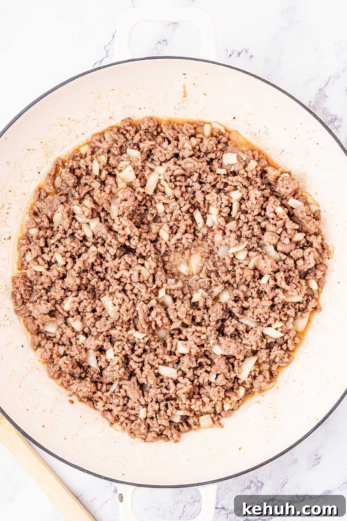 Savory Sloppy Joe Sliders Recipe for Game Day and Parties 14 A large white pan filled with cooked, seasoned ground meat and diced onions sits on a marble countertop, ready to be transformed into delicious sloppy joes. A wooden spatula is partially visible to the left.