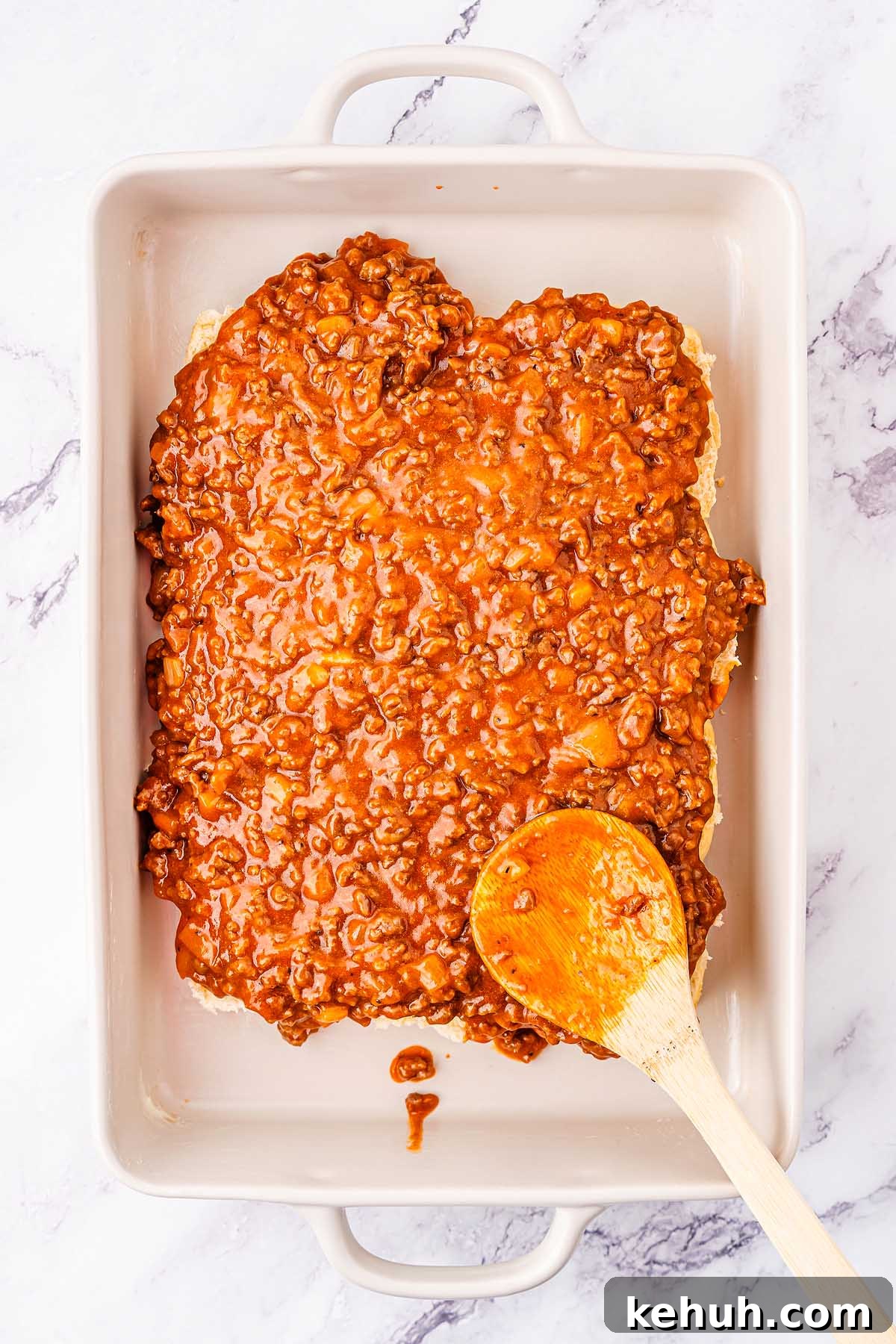 Savory Sloppy Joe Sliders Recipe for Game Day and Parties 17 A rectangular baking dish filled with a thick layer of ground meat sauce, reminiscent of sloppy joes, spread with a wooden spoon placed inside the dish. The surface underneath is white with subtle gray veins.