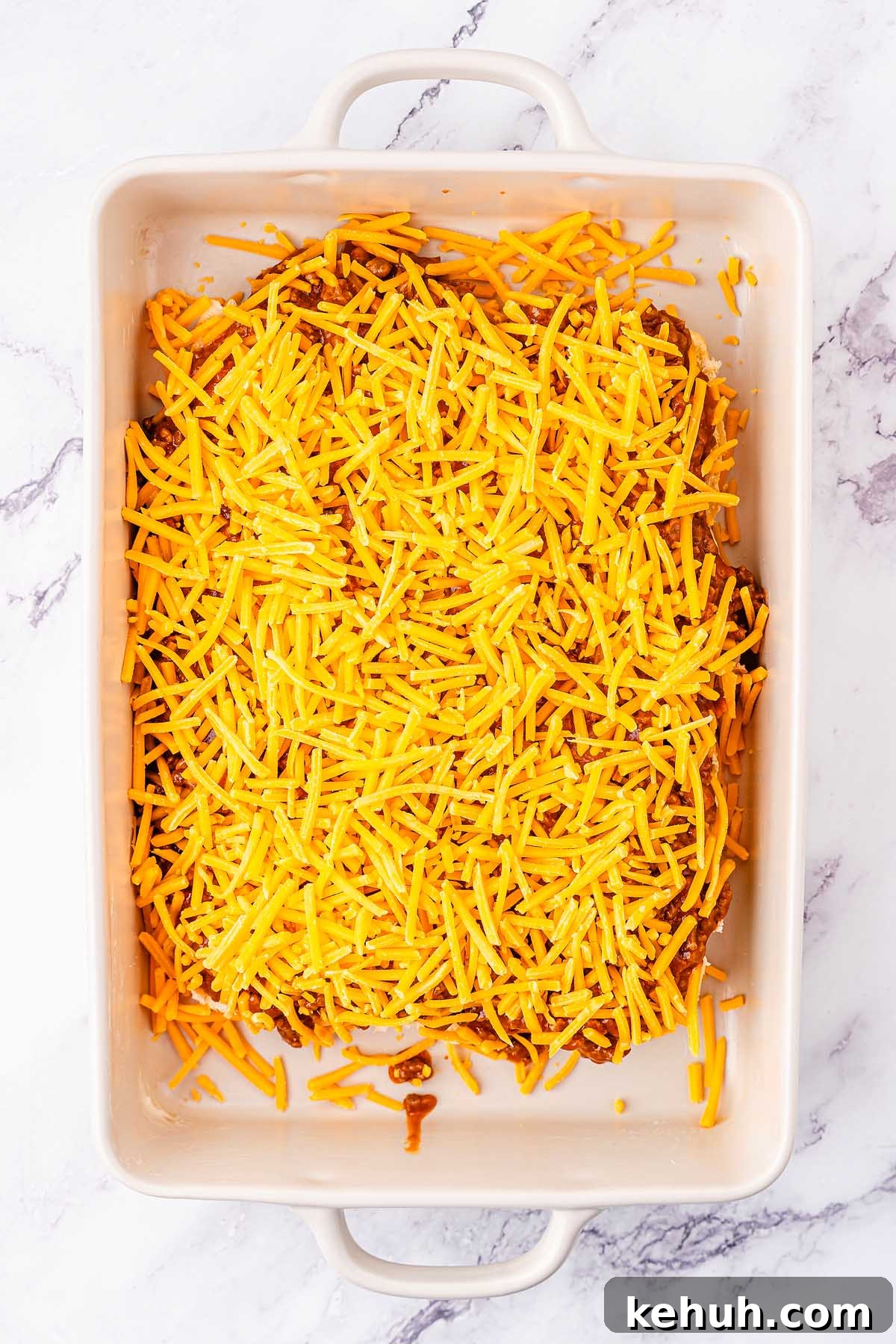 Savory Sloppy Joe Sliders Recipe for Game Day and Parties 18 A rectangular baking dish filled with shredded cheddar cheese atop a savory layer of sloppy joes, placed on a marble surface.