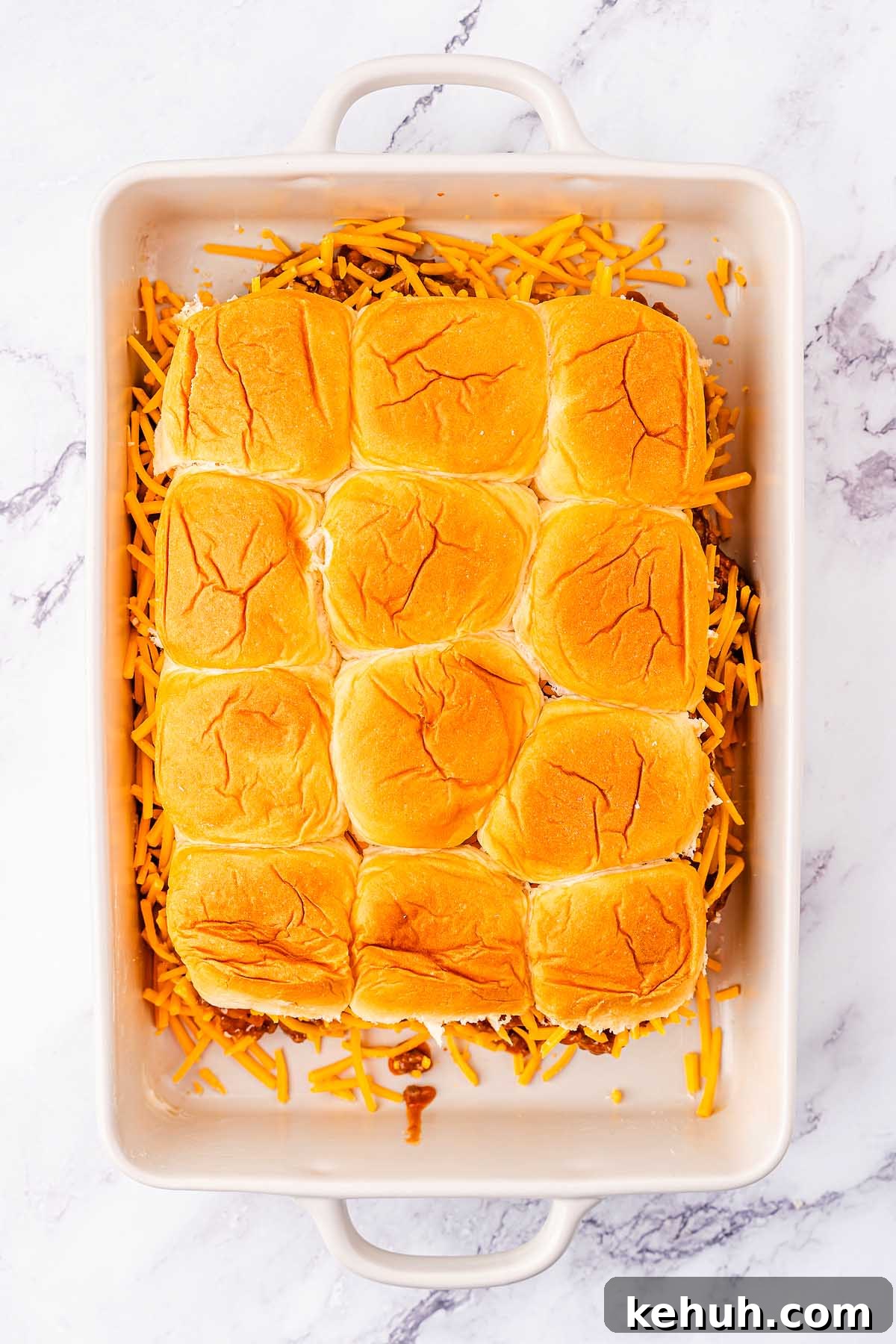Savory Sloppy Joe Sliders Recipe for Game Day and Parties 19 A white baking dish containing a partially cooked batch of 12 dinner rolls with shredded cheddar cheese around the edges on a marble countertop, perfect for transforming into sloppy joe sliders.