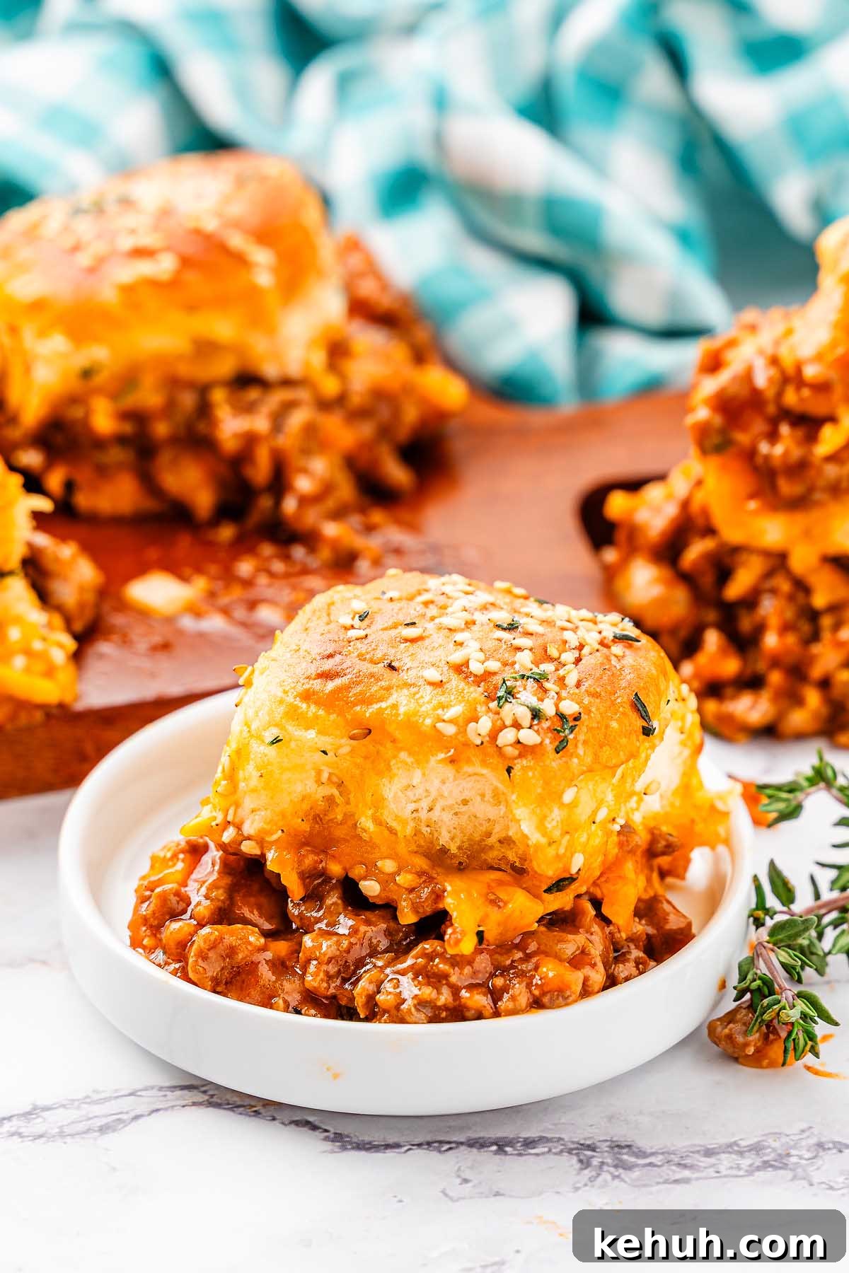 Savory Sloppy Joe Sliders Recipe for Game Day and Parties 3 A small white dish holds a sloppy joe slider with meat, cheese, and sauce, topped with a golden bun and garnished with herbs. More sliders and a blue checkered cloth are in the background.