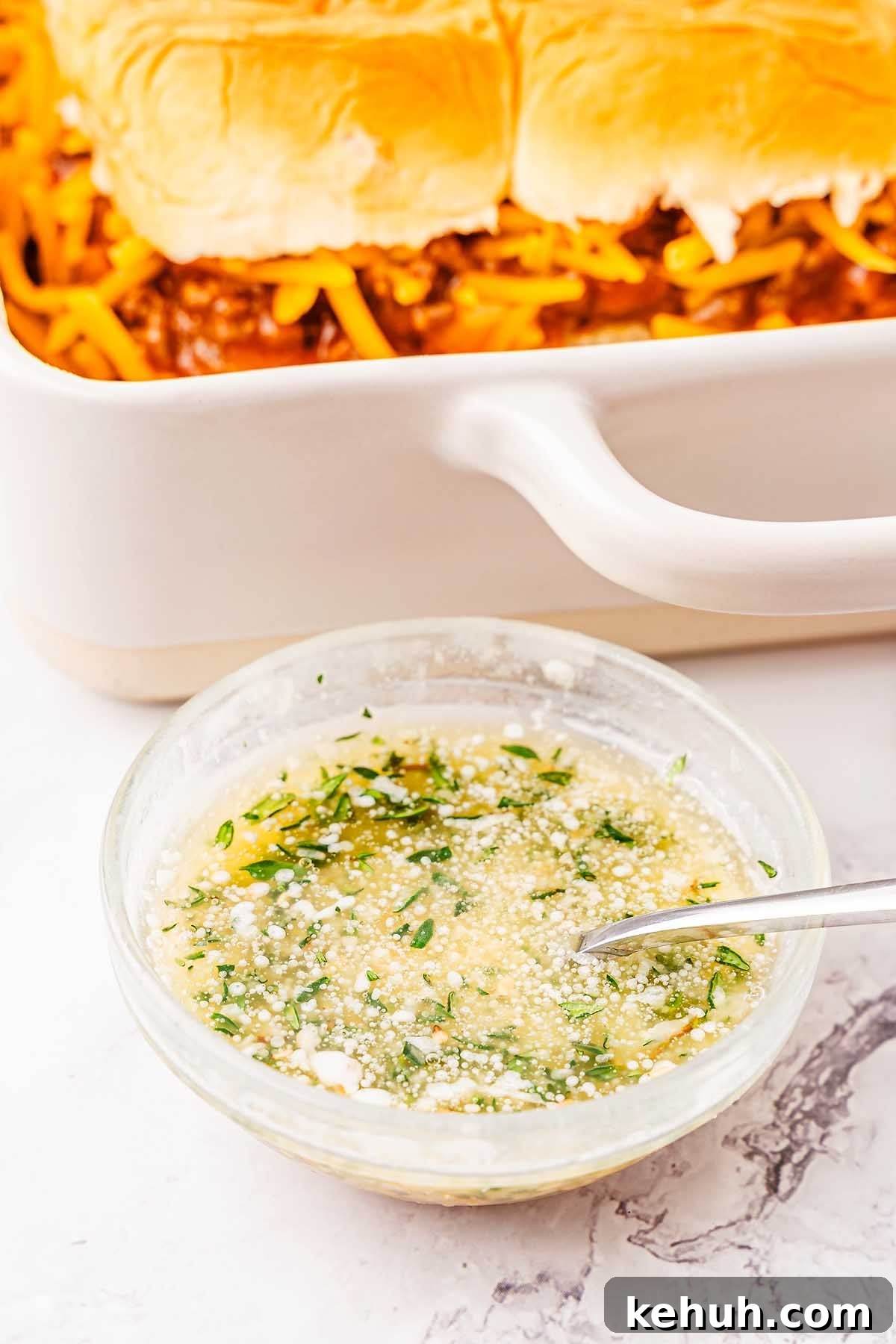 Savory Sloppy Joe Sliders Recipe for Game Day and Parties 20 A glass bowl with a herb and cheese mixture in front of a baking dish containing bread rolls, reminiscent of sloppy joe sliders, and what appears to be a cheesy filling. A spoon rests in the bowl.