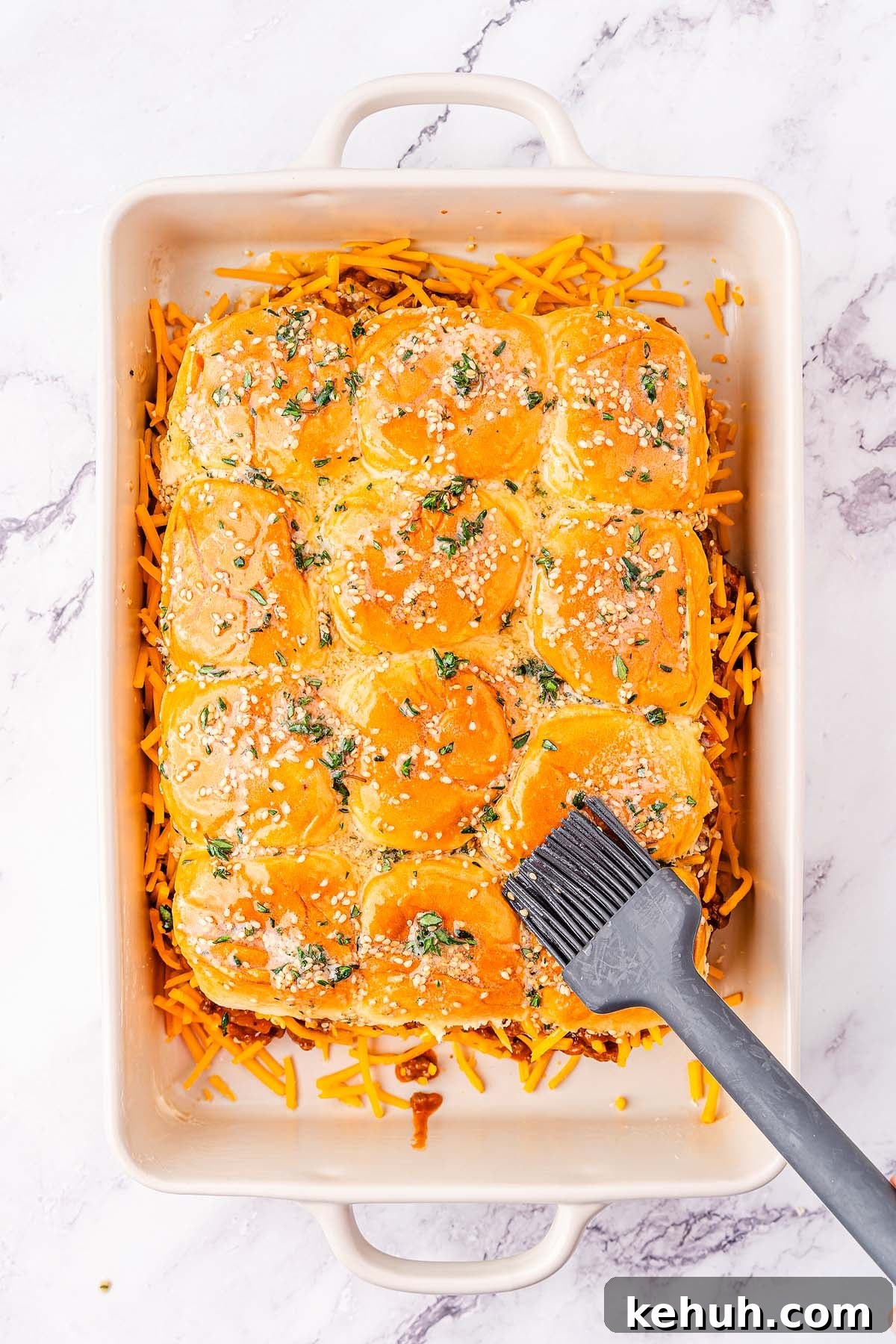 Savory Sloppy Joe Sliders Recipe for Game Day and Parties 21 A baking dish filled with sloppy joes sliders topped with sesame seeds and herbs. A brush is applying melted butter to the tops of the buns while shredded cheese is scattered around the edges.