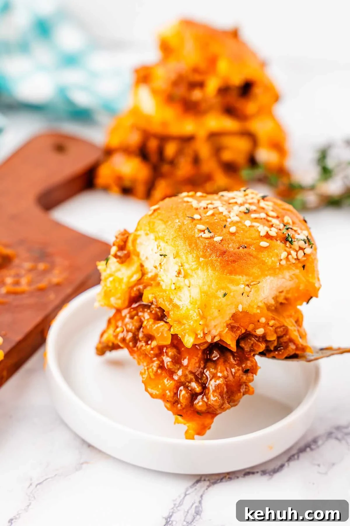 Savory Sloppy Joe Sliders Recipe for Game Day and Parties 4 A close-up of a cheesy, sesame-topped sloppy joe's slider filled with ground meat and sauce on a small white plate. More sliders are visible in the background on a wooden cutting board.