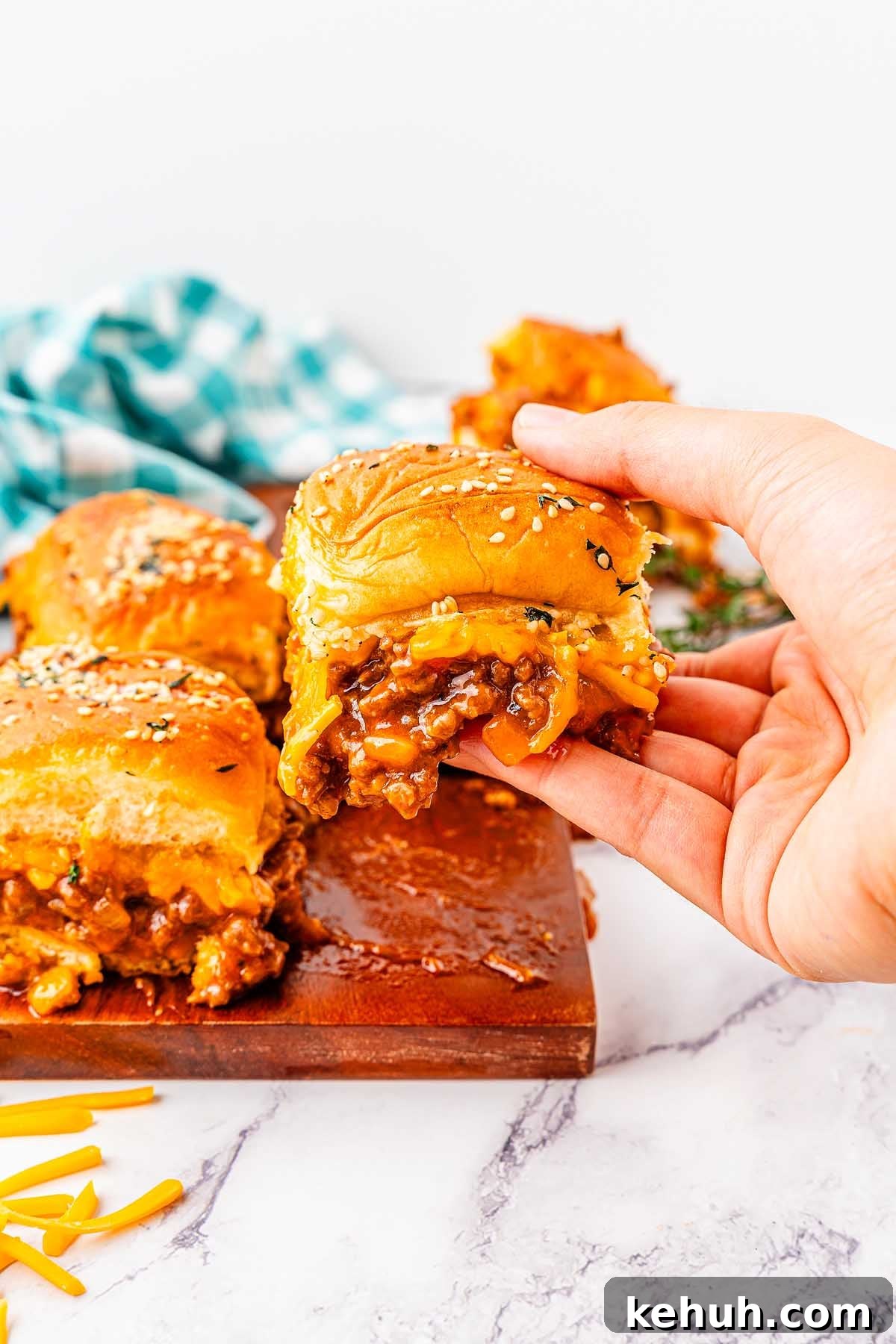 Savory Sloppy Joe Sliders Recipe for Game Day and Parties 7 A hand holding a sloppy joe sandwich. Shredded cheese pieces are in the background on a wooden board and a marble surface.