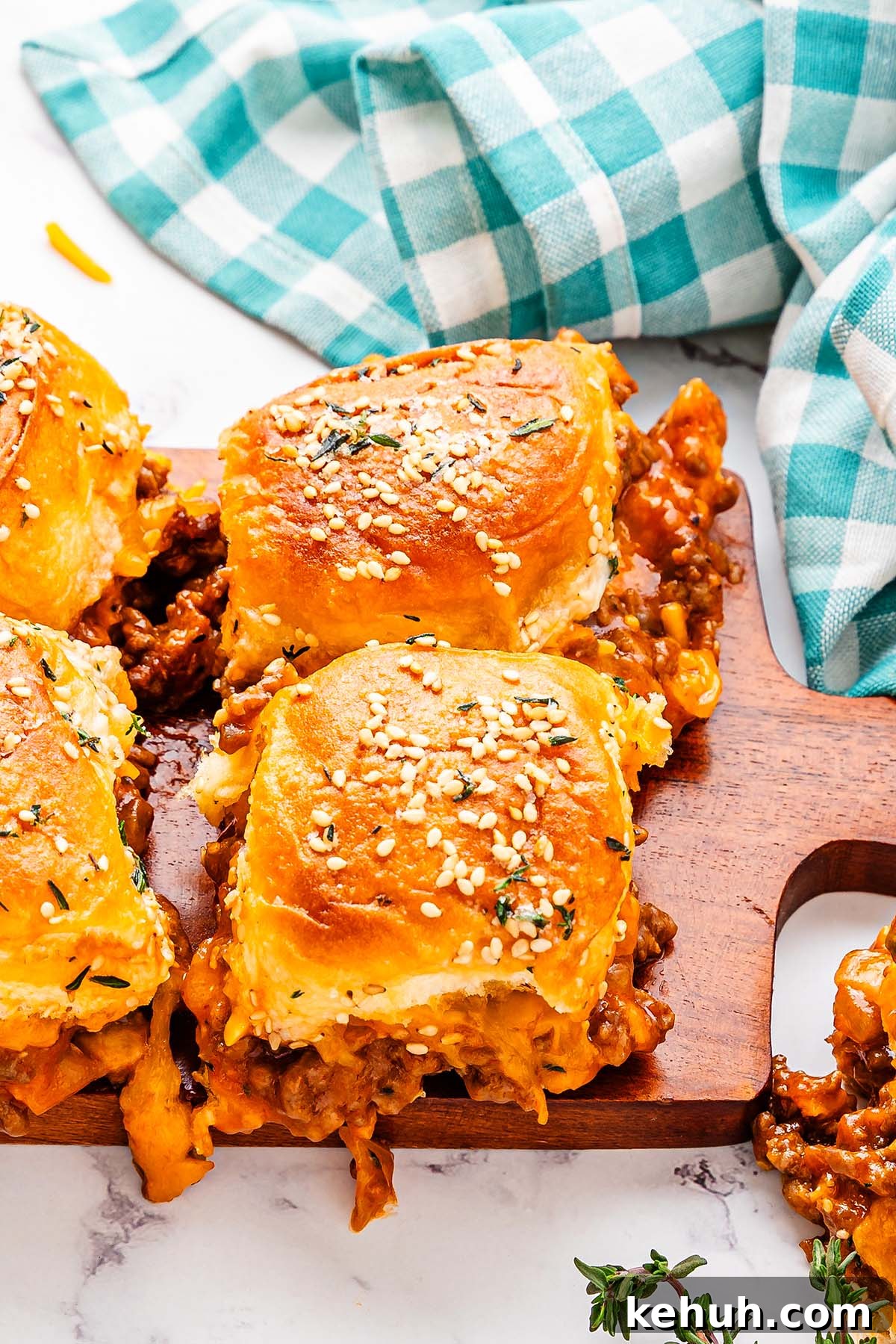 Savory Sloppy Joe Sliders Recipe for Game Day and Parties 9 Sliders with sesame seed buns filled with a savory sloppy joes and cheese mixture are placed on a wooden board next to a blue and white checkered cloth.
