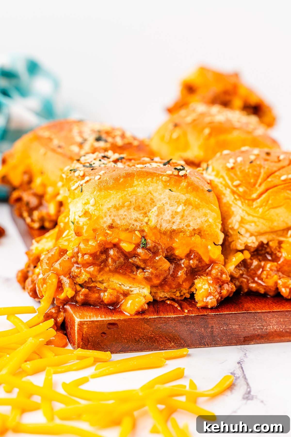Savory Sloppy Joe Sliders Recipe for Game Day and Parties 10 Close-up of three cheesy sloppy joe sliders with sesame seed buns on a wooden board. Shredded cheese is scattered around the sliders.