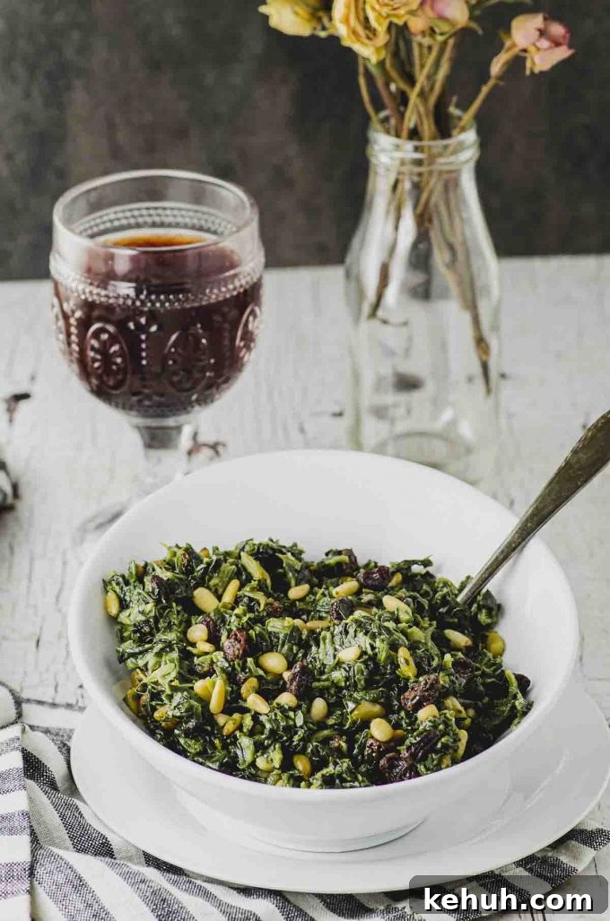 Side view of a white bowl filled with spinach with pine nuts and a raisins with a glass of red wine in the background.