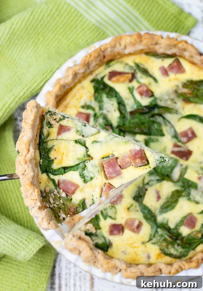 Spinach, Ham & Cheese Quiche Recipe for Brunch and Weeknights 2 A piece of ham and cheese quiche is lifted above the pie with a serving utensil. A green napkin rests beside it.