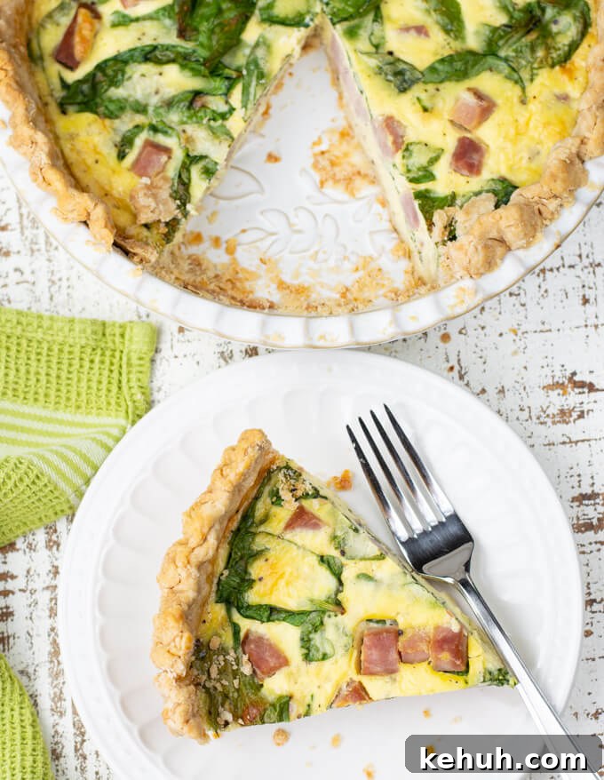 Spinach, Ham & Cheese Quiche Recipe for Brunch and Weeknights 8 A piece of ham and cheese quiche sits on a white plate with a fork. The pie pan of quiche sits next to it.