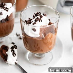 Parfait goblets filled with chocolate mousse, whipped cream, and chocolate curls with a spoonful laying next to one on a white marble slab and white textured background.