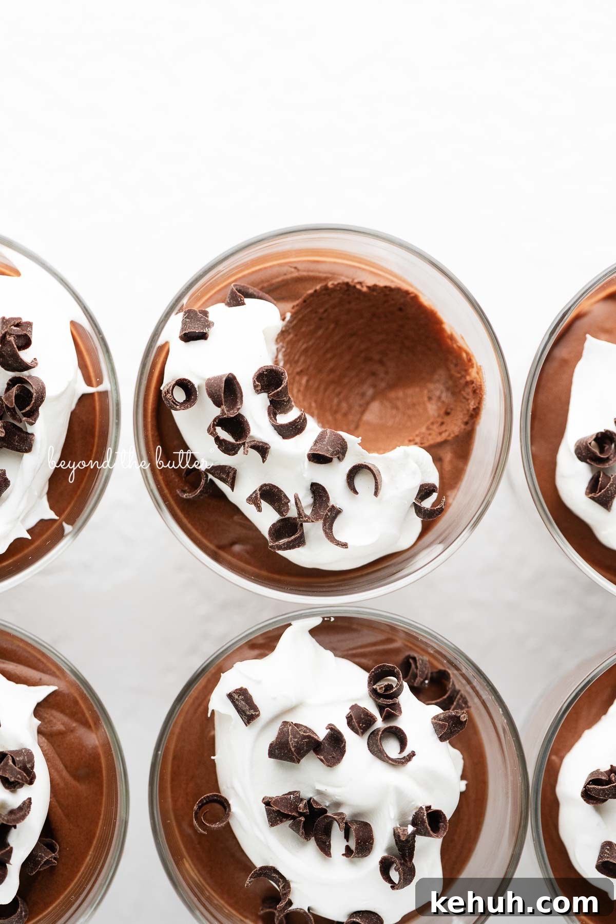 Six parfait goblets filled with chocolate mousse, whipped cream, and chocolate curls with a spoonful removed from the top center goblet on a white textured background.