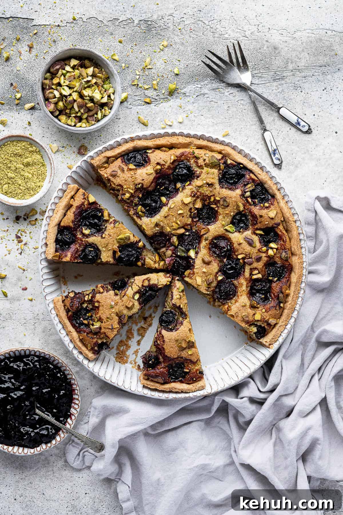 Vegan Pistachio and Cherry Frangipane Tart Recipe 7 The sliced pistachio cherry frangipane tart on a grey surface with bowls of jam and pistachios.