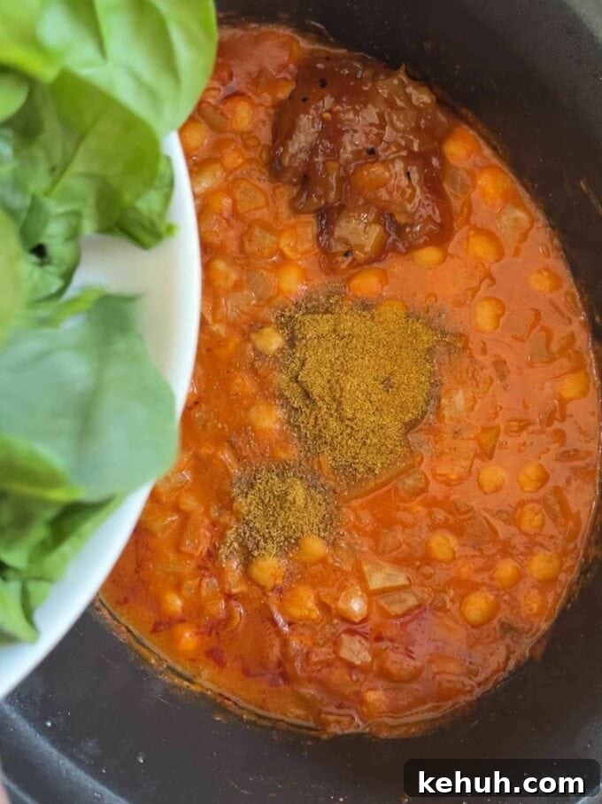 A slow cooker pan filled with chickpeas in a sauce, with spices on top and spinach about to the tipped in.