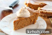 A slice of pumpkin pie with a dollop of whipping cream on top sitting on a small dessert plate. Faded in the background is a pie server and a pumpkin pie with a slice cut out of it.