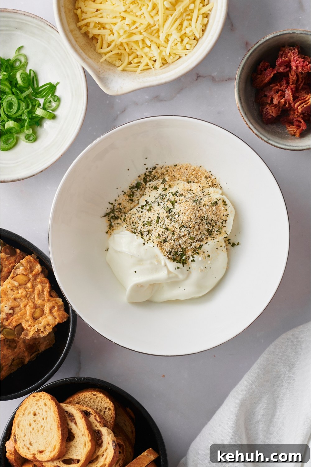 Ranch Bacon Cheddar Crack Dip Recipe for Parties 5 White mixing bowl with seasoning blend and sour cream, surrounded by bowls of shredded cheese, green onion, bacon bits, bread slices, and tortilla chips.