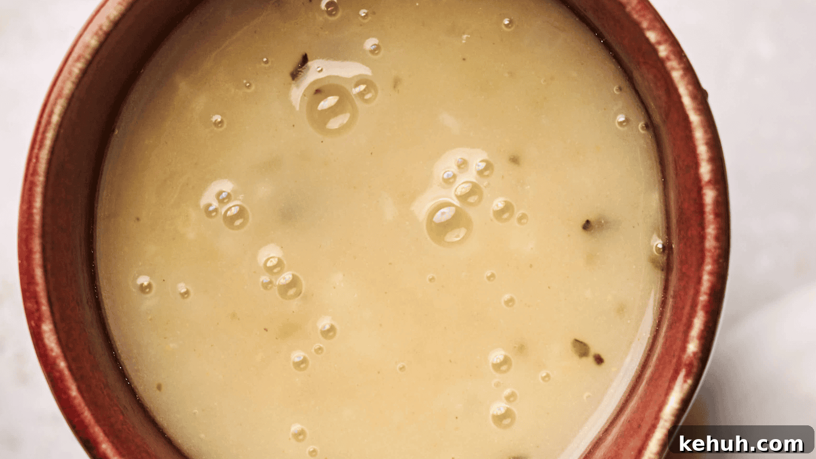 31 Homemade Thanksgiving Sides That Outshine Store-Bought 26 Close-up of a bowl filled with creamy turkey gravy soup, featuring small bubbles and herbs on the surface. The bowl has a reddish-brown rim.