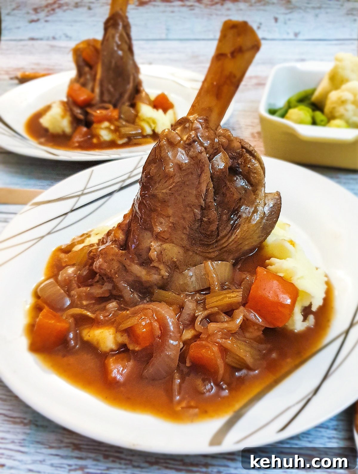 A lamb shank on a pile of mashed potatoes covered with red wine gravy and vegetables.