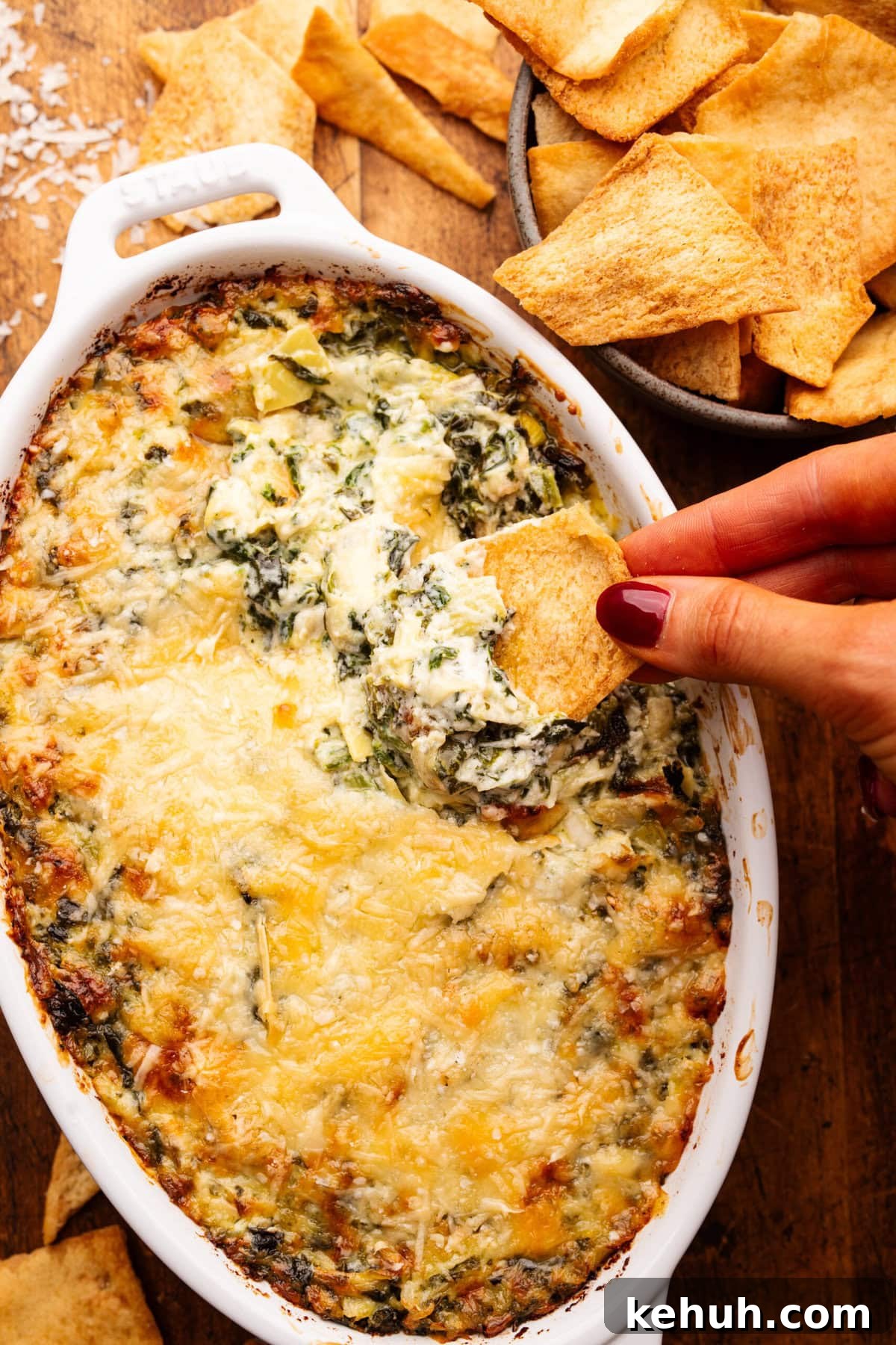 A close up of a hand dipping a pita chip into a white oval dish with baked artichoke dip in it. Around the dip is a bowl of pita chips.