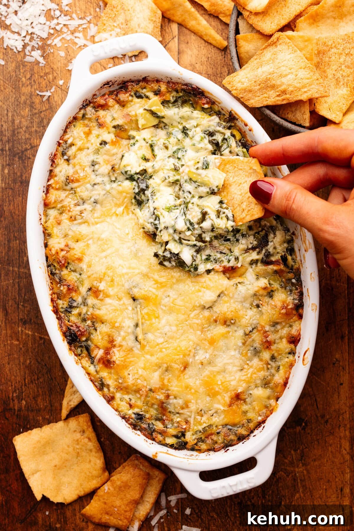 A hand dipping a pita chip into a white oval dish with baked artichoke dip in it. Around the dip is a bowl of pita chips. 