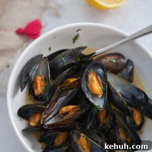 Mussels in Rich Lobster Broth: Savory Seafood Stew Recipe 7 black opened mussels with a spoon in a white bowl with a lemon on the side.