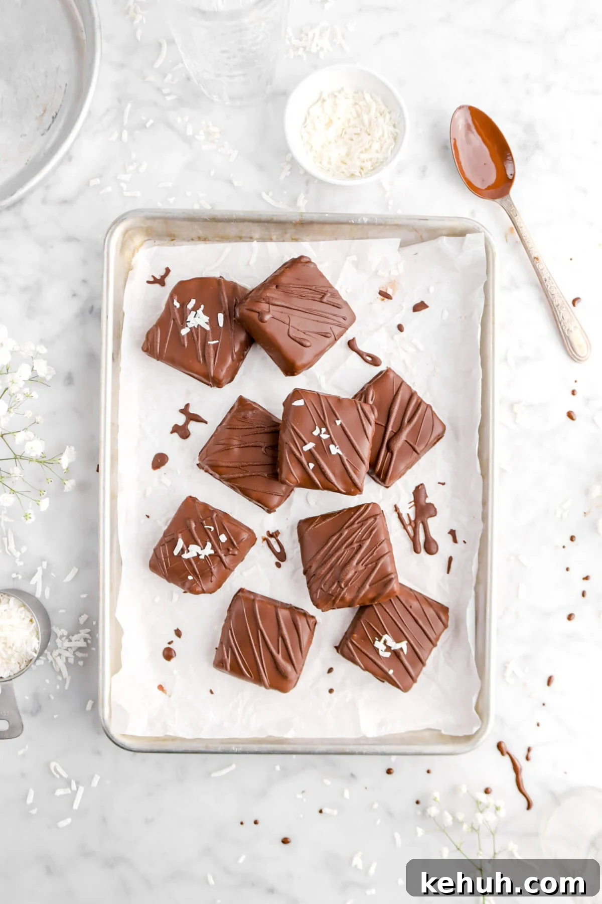 Coconut Cream-Filled Chocolate Truffles: Decadent Homemade Recipe 18 overhead shot of nine coconut chocolates on sheet pan with spoonful of melted chocolate, coconut flakes, and white flowers around on marble surface