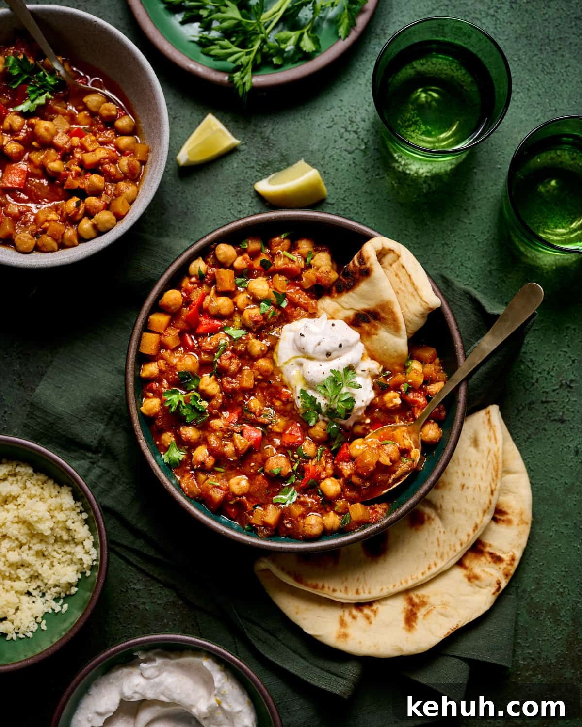 Bowl of tagine with pita bread and yogurt next to a bowl of couscous on a dark green table.