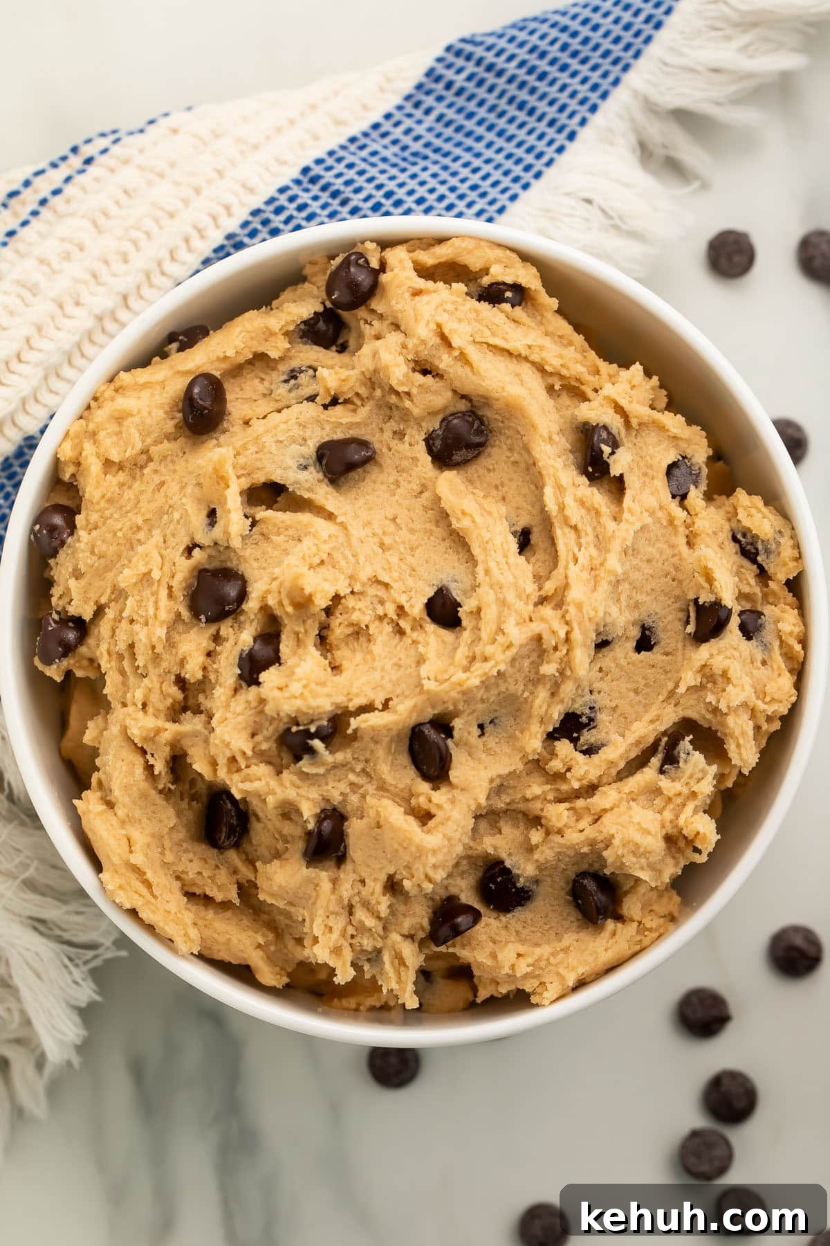 Vegan Cookie Dough Recipes: Safe-to-Eat & No-Bake Options 4 A bowl of vegan cookie dough rests on a kitchen counter, surrounded by loose chocolate chips and a blue and white striped kitchen towel.
