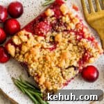 Top down shot of a cranberry bar on a plate with a fork, some raw cranberries and a few pieces of rosemary.
