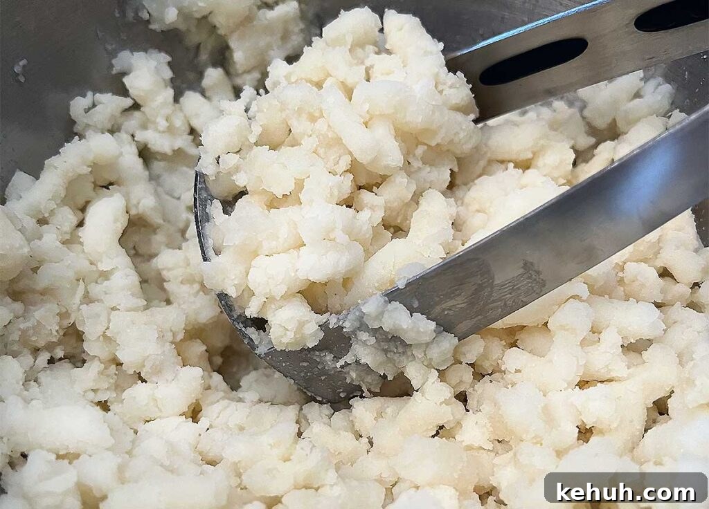 Cooked potatoes being mashed with a large silver potato masher.
