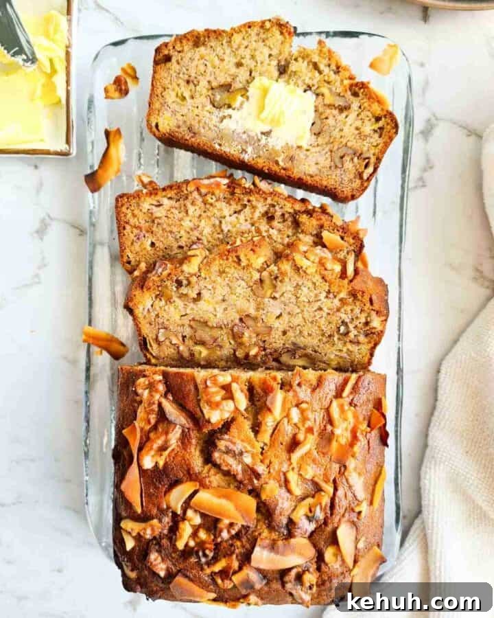 A loaf of banana bread with half thickly sliced on a glass plate. One slice with butter spread on top.