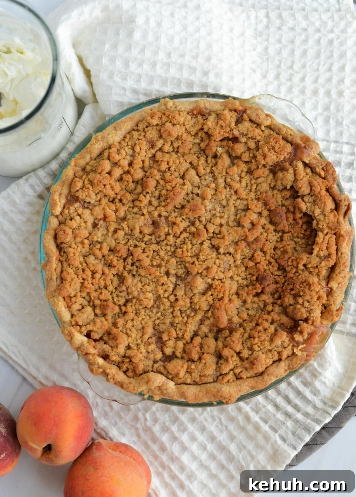 Sourdough Peach Crumb Pie Recipe with Flaky Rustic Crust 11 A baked peach pie sits on a kitchen towel next to a jar of fresh whipped cream and several whole peaches.