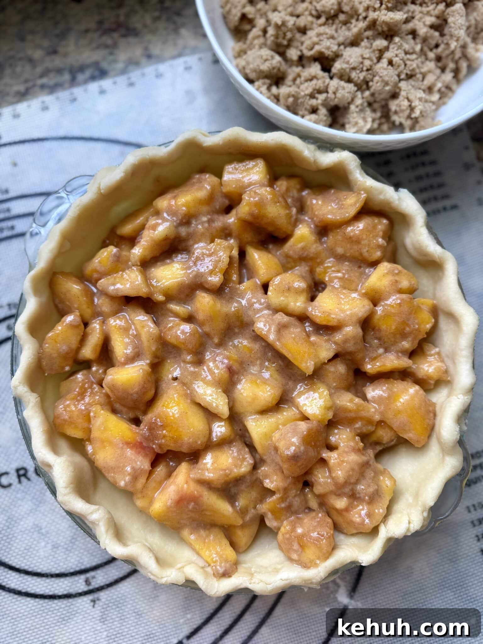 Sourdough Peach Crumb Pie Recipe with Flaky Rustic Crust 10 Peach pie with a sourdough crust sitting on a pastry mat filled with peaches and a crumb topping sitting in a bowl next to the pie plate.