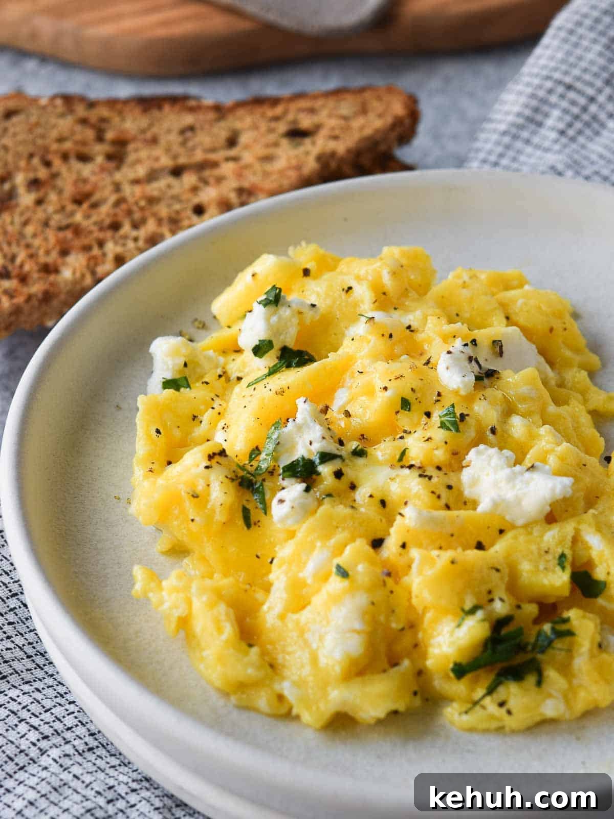 Creamy Goat Cheese Scrambled Eggs with Herbs and Cherry Tomatoes 10 Scrambled eggs with goat cheese on a white plate with toast in the background.