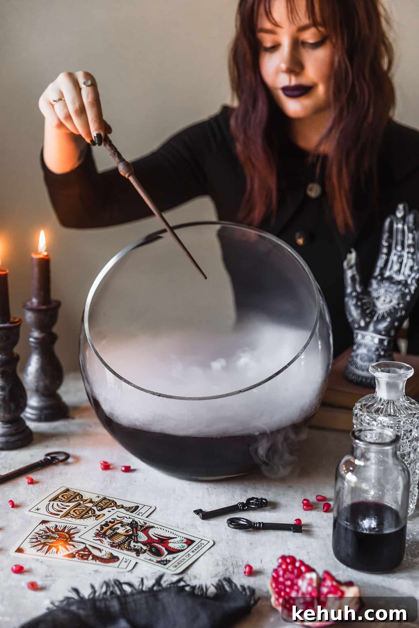 Spooky Witches Brew Punch Recipe for Halloween Parties 6 A woman in a black dress waving a wand over a bowl of witches brew cocktail punch next to potion bottles, vintage keys, and tarot cards on a grey background.