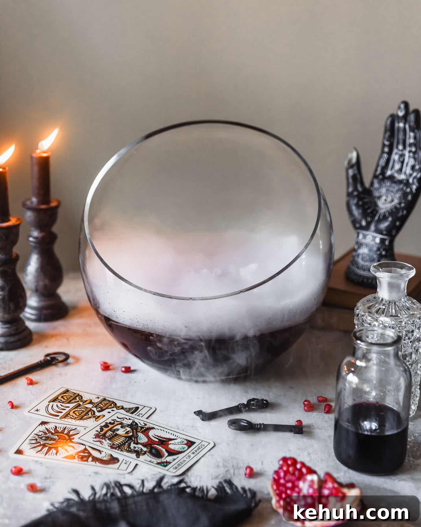 Spooky Witches Brew Punch Recipe for Halloween Parties 7 A bowl with a purple Halloween drink on a white counter next to lit black candles, bottles of alcohol, Halloween decorations, and tarot cards.