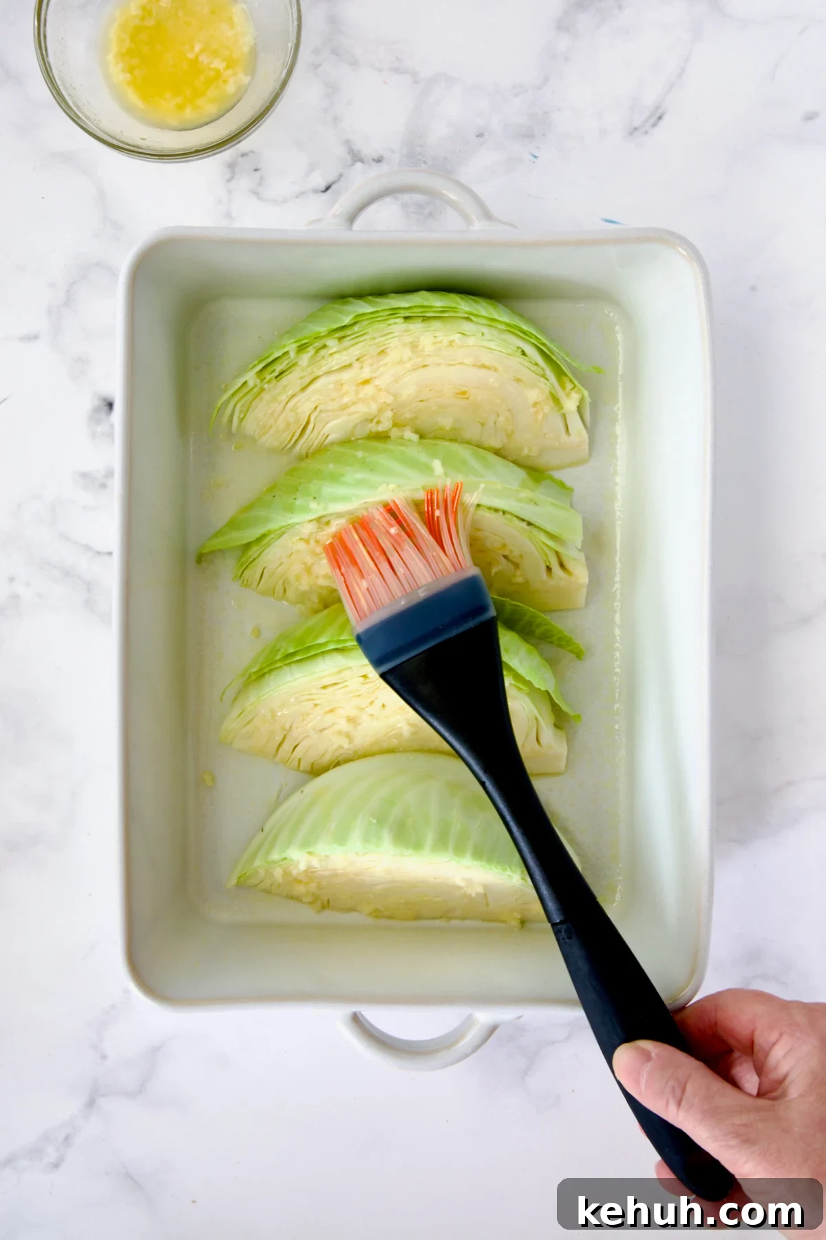 Four wedges of cabbage in white baking dish being brushed with butter and garlic.