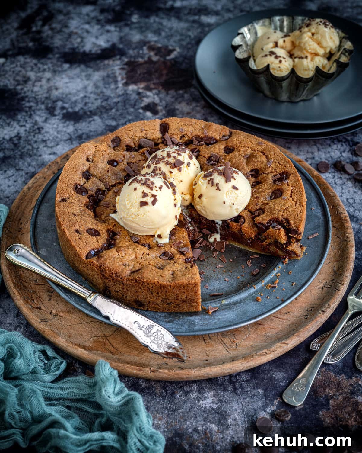 Giant Chocolate Chip Cookie Recipe — Air Fryer or Oven 3 A giant chocolate chip cookie cake is topped with vanilla ice cream scoops and chocolate shavings, served on a round metal tray with a knife, fork, and extra ice cream in a bowl on a textured dark surface.