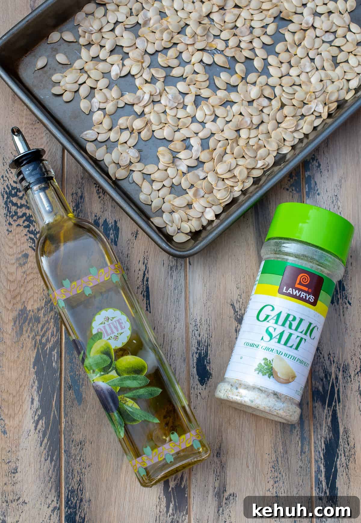 A top down shot of acorn squash seeds on a baking sheet, a glass bottle of olive oil, and a container of garlic salt on a brown board.