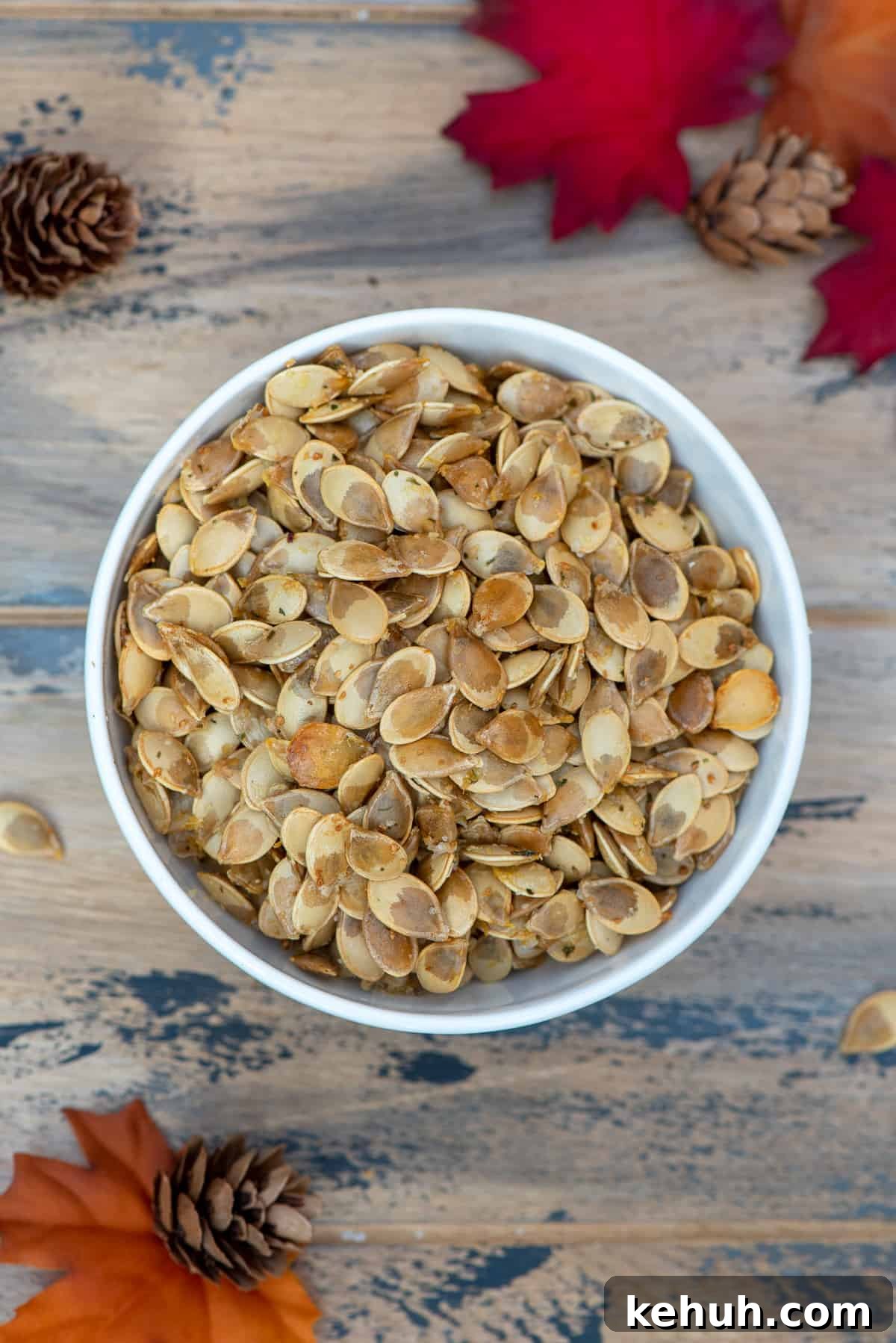 A top down shot of a bowl of roasted acorn squash seeds on a brown ball with fall leaves and pine cones.