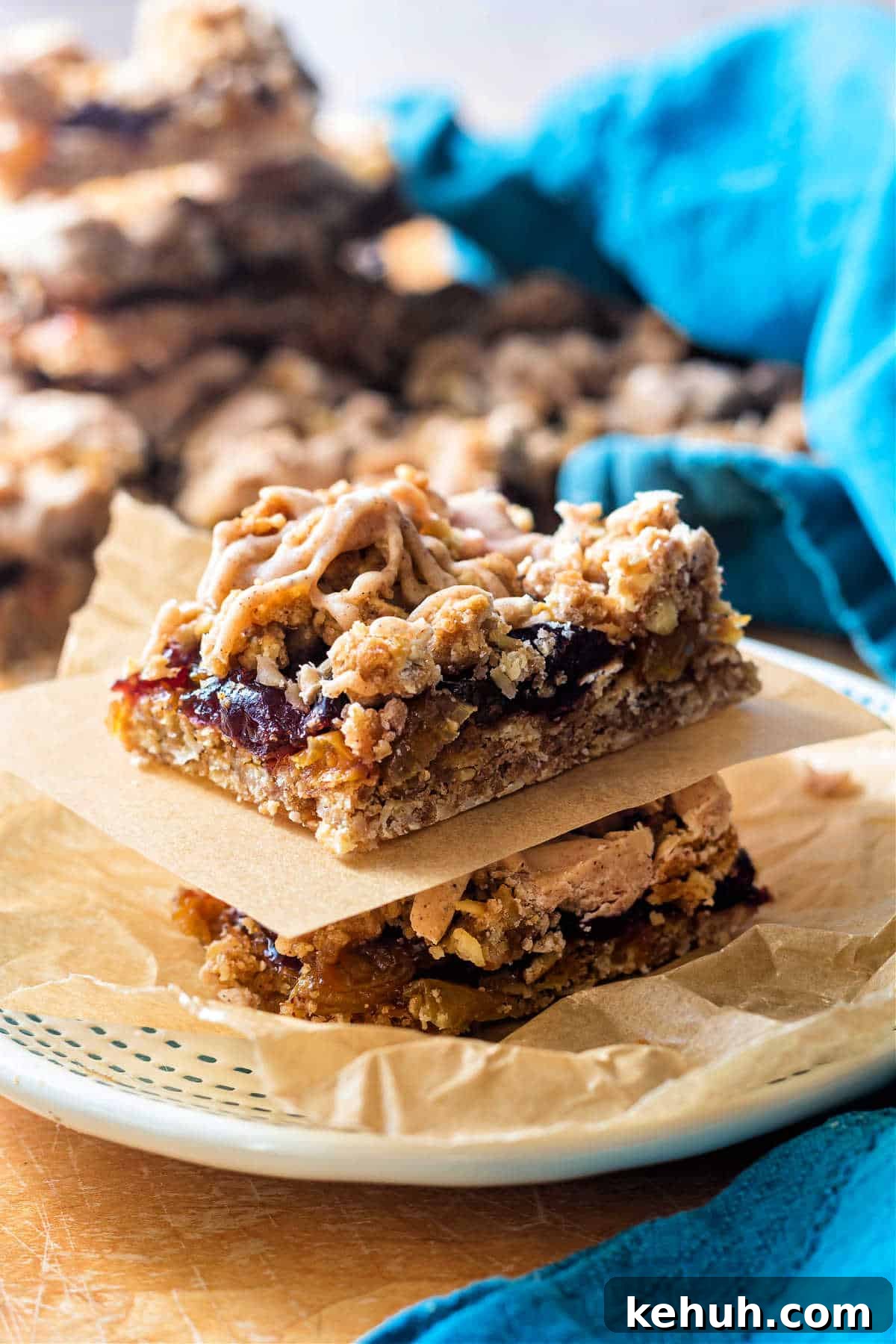 Grandma’s Oatmeal Raisin Bars Recipe for Chewy, Home-Baked Treats 2 A vertical image of two stacked cranberry raisin bars on a small plate, separated by a small rectangle of parchment. There are more raisin bars in the background as well as a turquoise-colred cloth napkin.