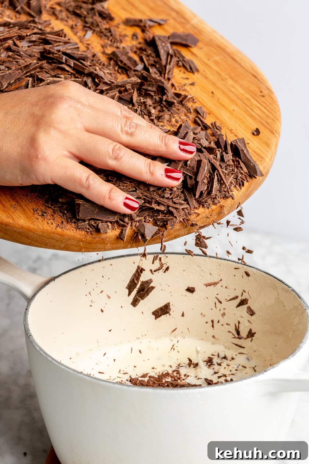 220824_Bourbon Pecan Truffles-05 Hand adding shaved chocolate into a white sauce pan with heavy cream from a wooden cutting board.
